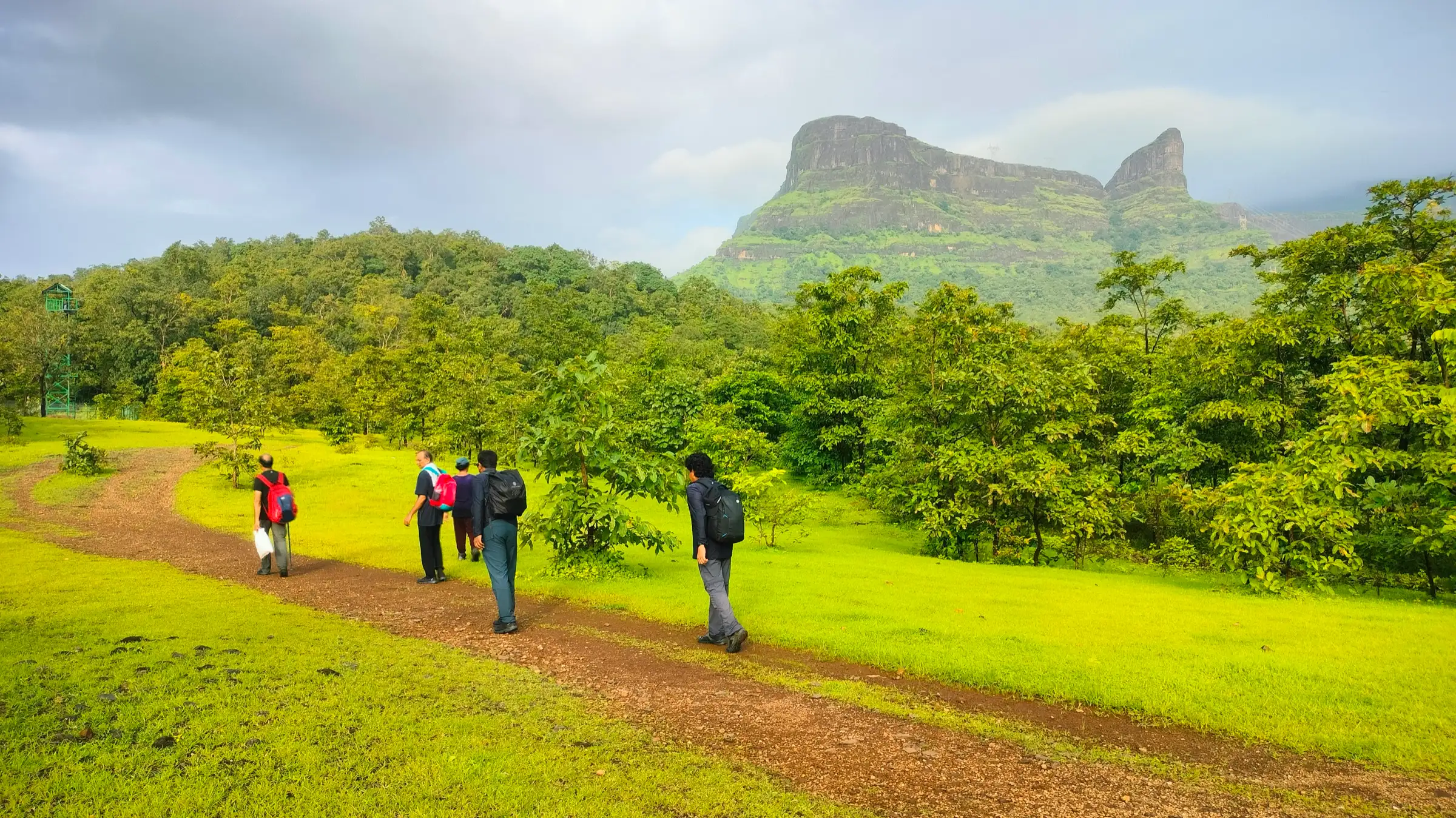 Trekkers walking a dirt trail through green monsoon meadow towards the flat-topped Naneghat cliffs and the Nanacha Angatha pinnacle