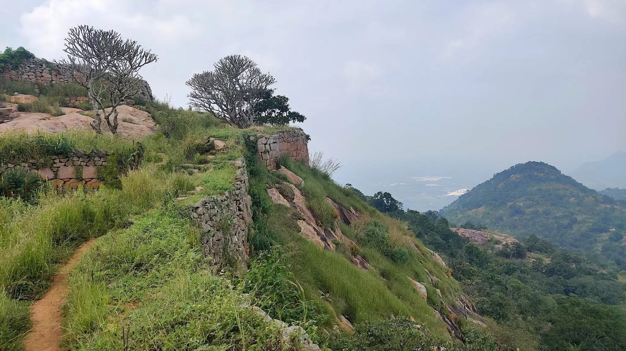 Dirt path along the ruined stone fort walls on top of Makalidurga, with a green hill and hazy plains beyond