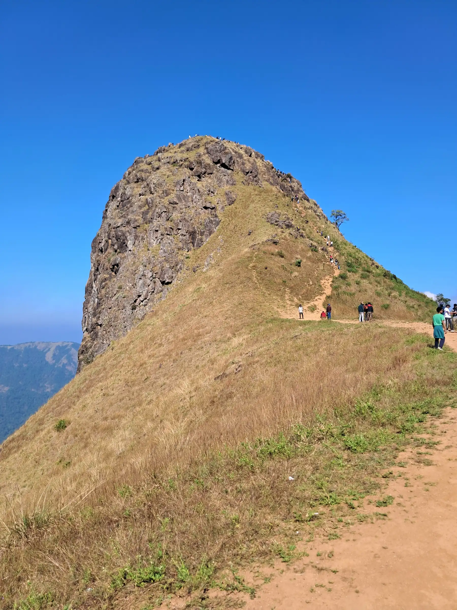 Trekkers on the dry grassy ridge below the rock hump of Ettina Bhuja under a blue sky