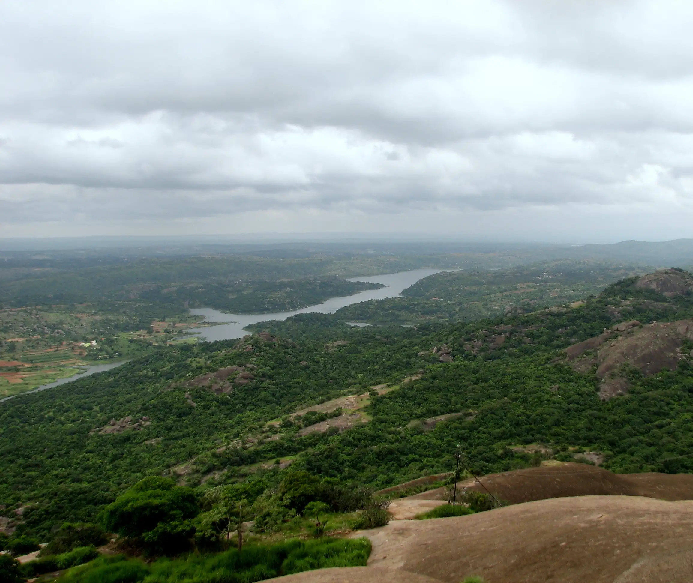 View from halfway up the bare granite of Savandurga over green forest to the winding Manchanabele reservoir under grey clouds