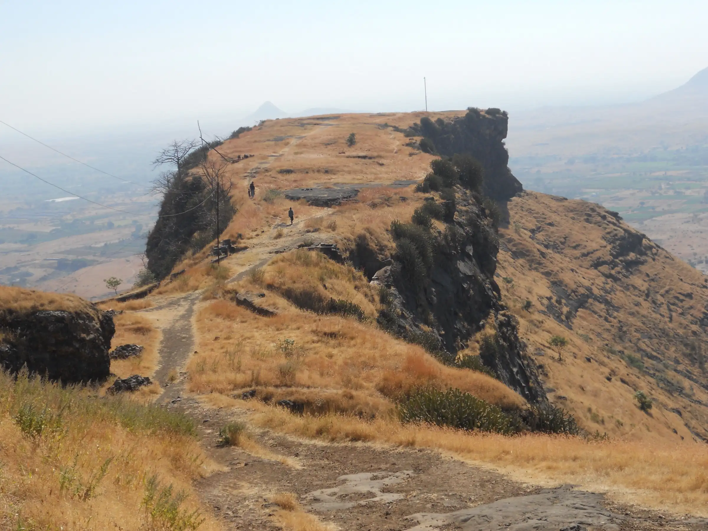 A dirt trail across the dry grassy top of Ramshej Fort, with walkers and a rock bastion above the hazy Nashik plains