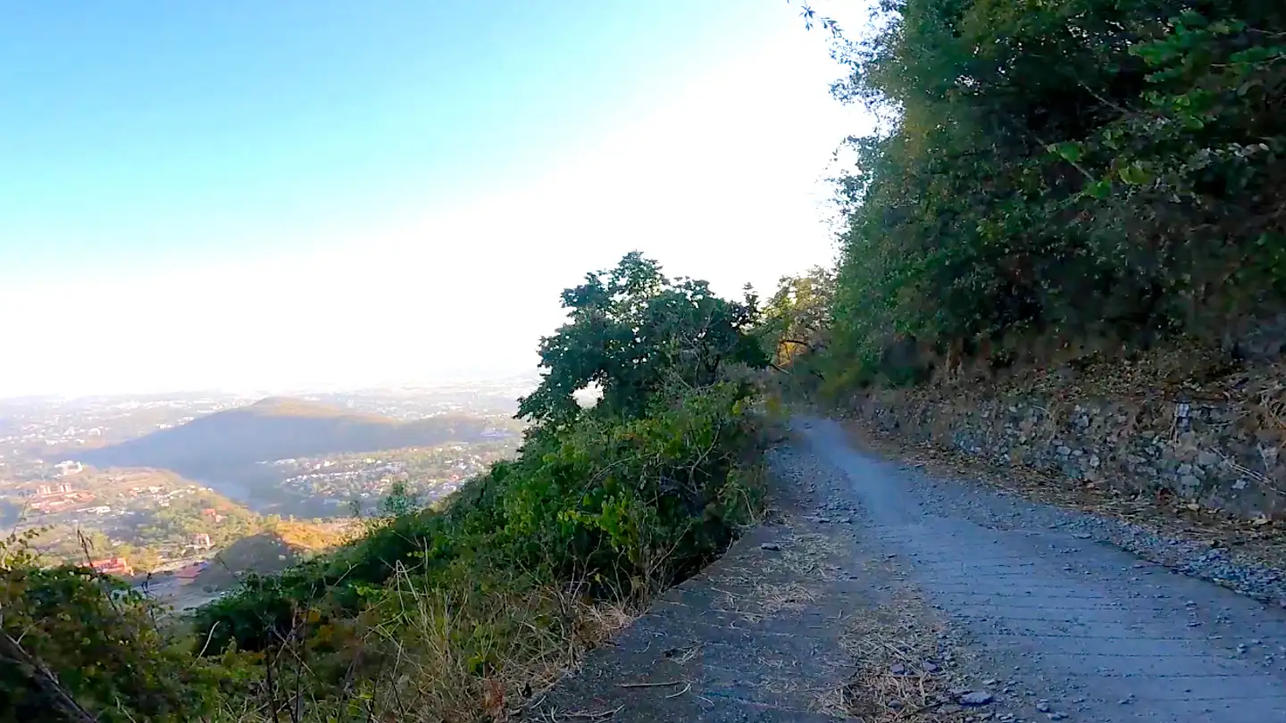 Narrow paved section of the Kipling Trail cut into a bushy hillside, with the Doon Valley and a hill far below