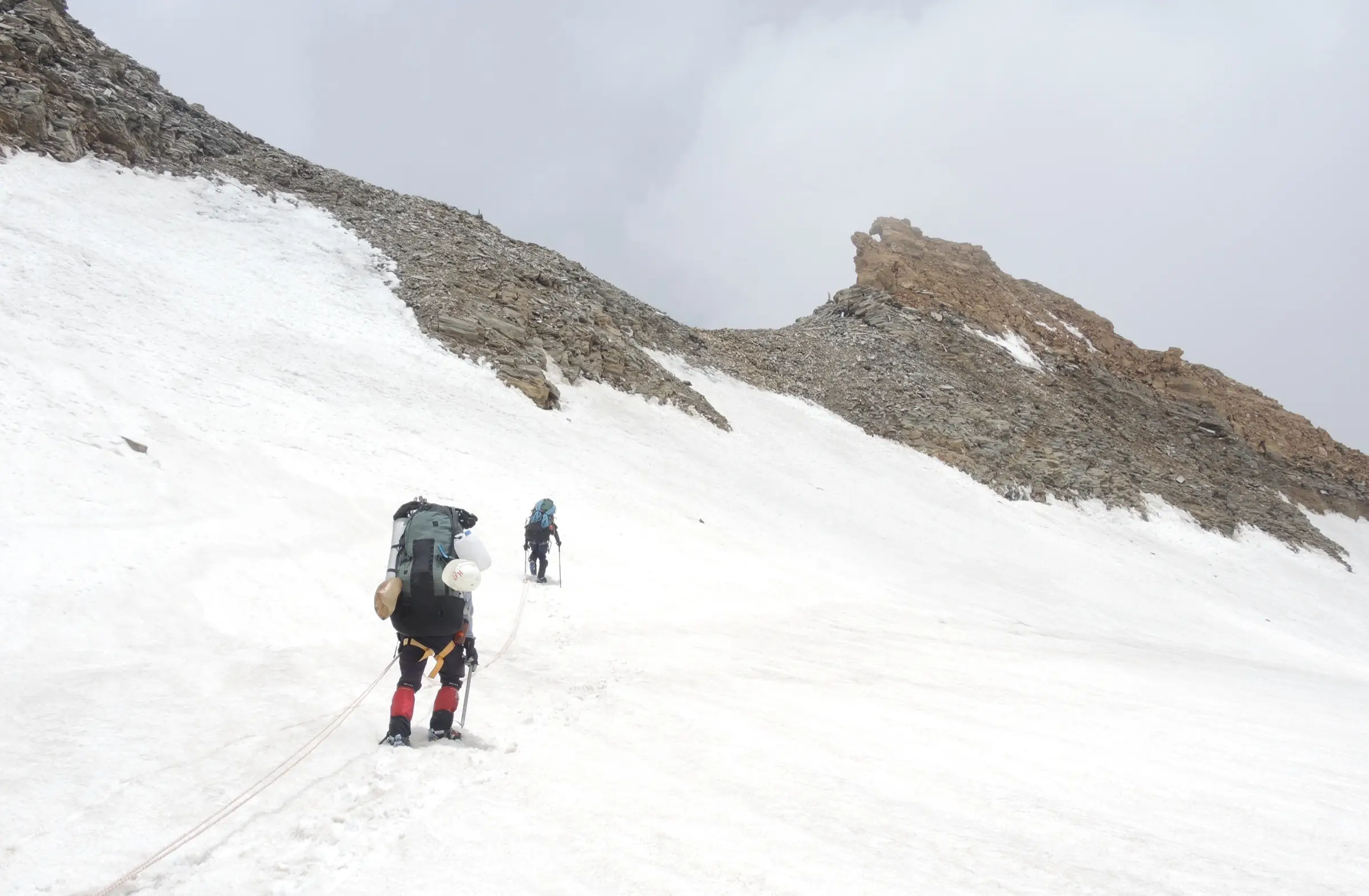 Two roped climbers with heavy packs walking up a snowfield towards a rocky ridge on Traill's Pass under grey cloud