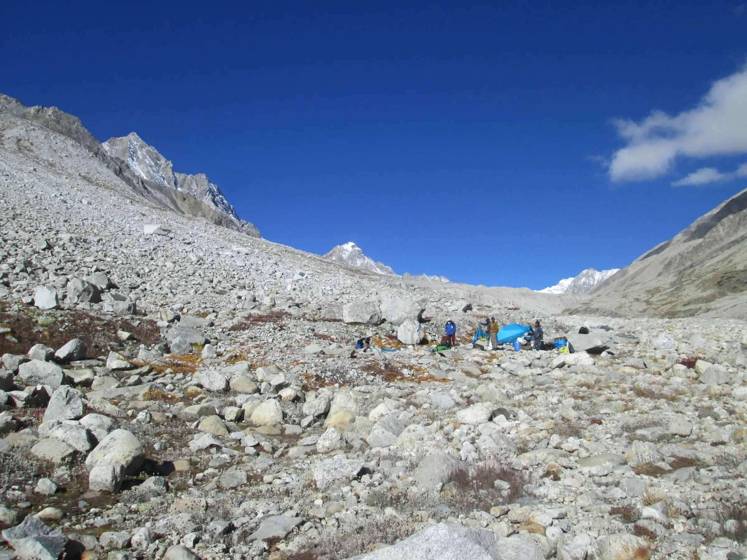Trekkers and a blue tarp at a camp on a vast boulder moraine below rocky snow peaks on the Kalindi Khal route