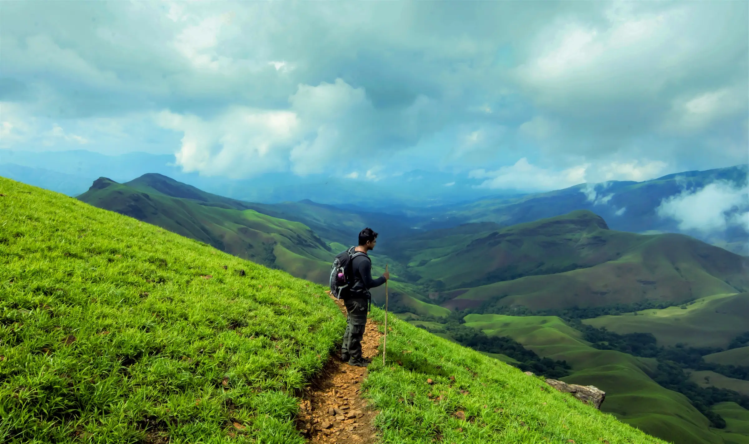Trekker on a narrow path across a steep green grass slope above the rolling hills of Kudremukh National Park