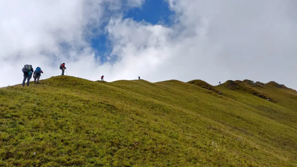 Trekkers with backpacks walking along a green meadow ridge at Gidara Bugyal
