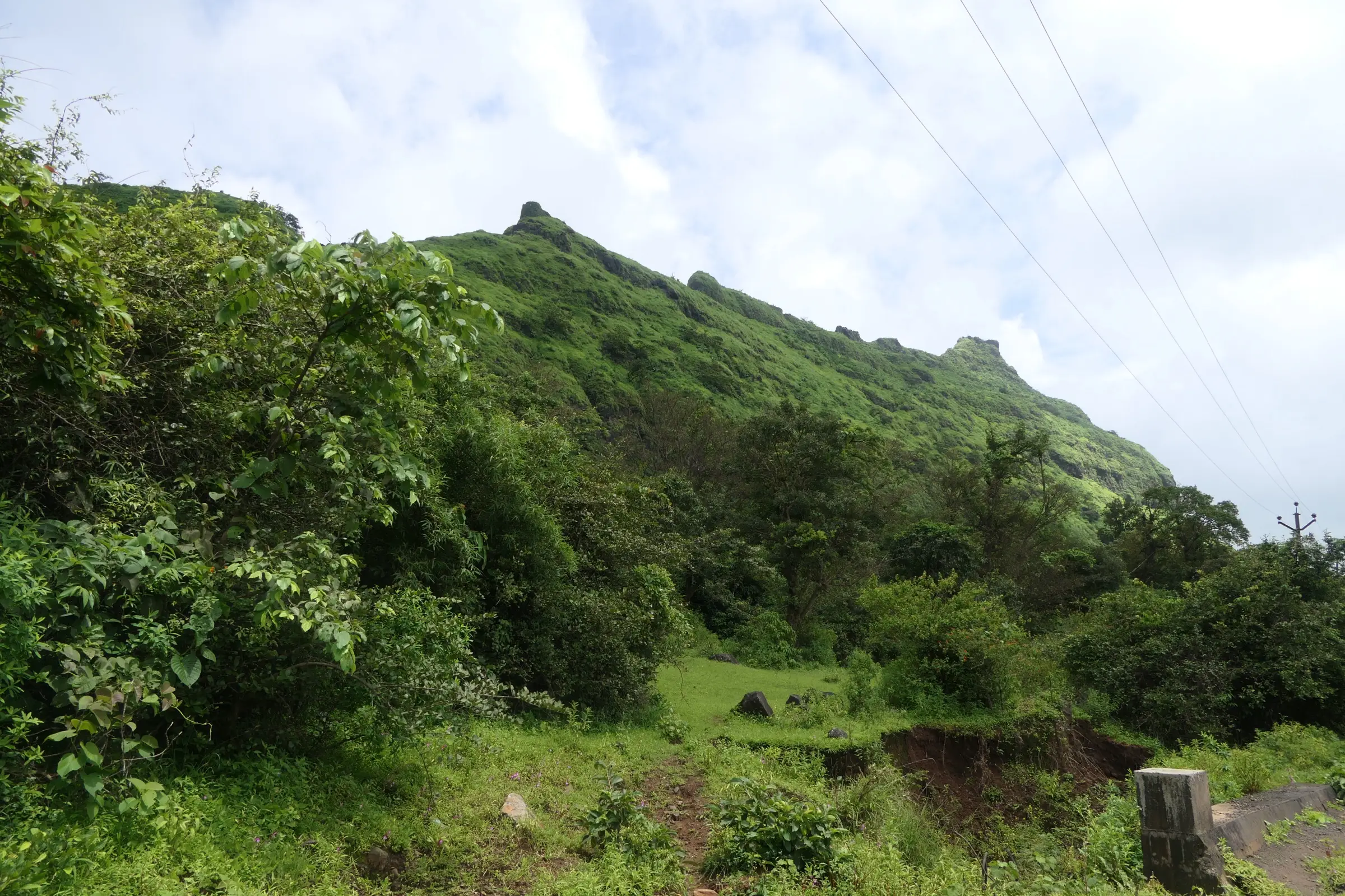 A footpath through green monsoon scrub at the base of Tung Fort, with the grassy ridge and bastions rising above