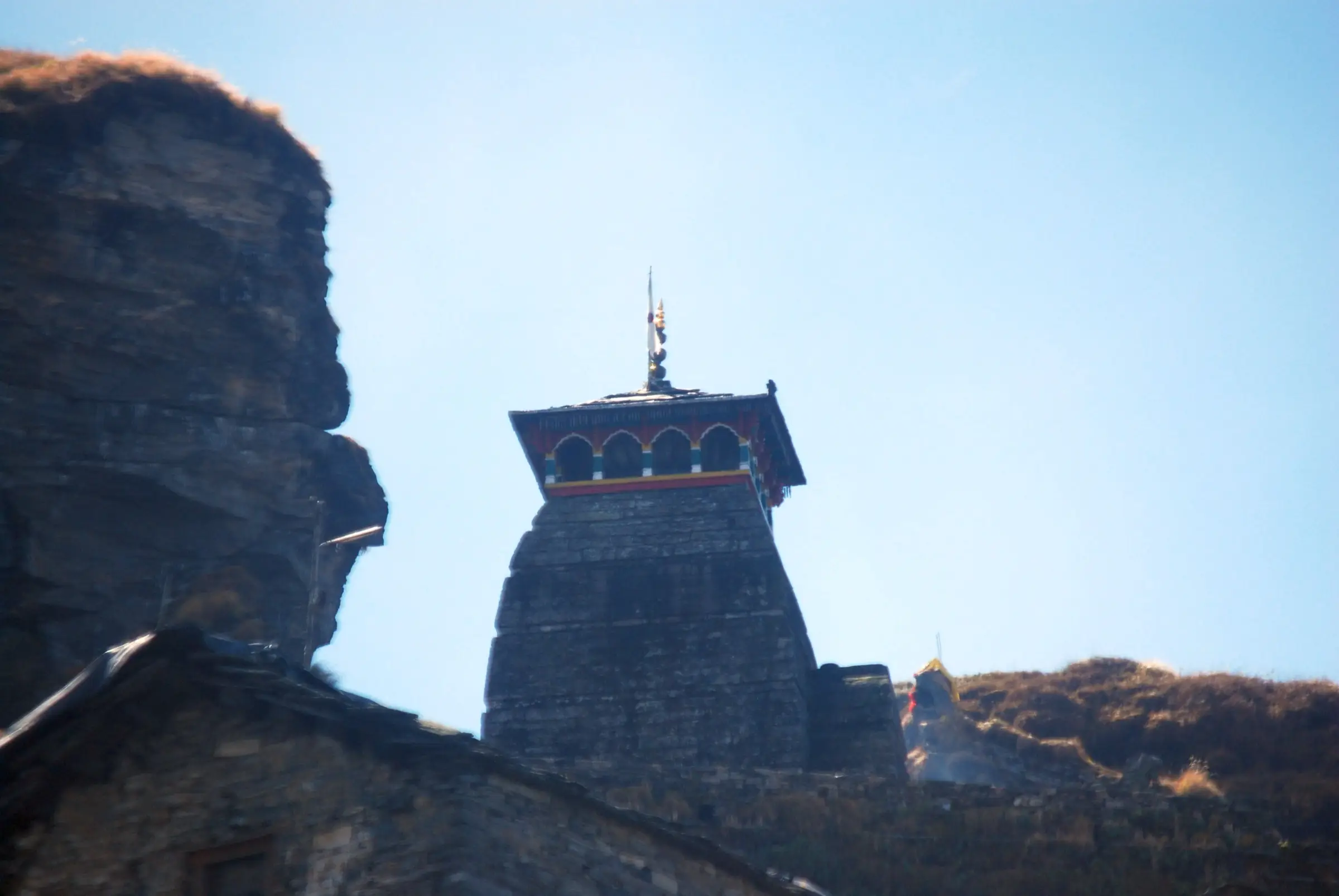 The stone shikhara of Tungnath temple with its painted wooden canopy, beside a rocky cliff under a clear sky