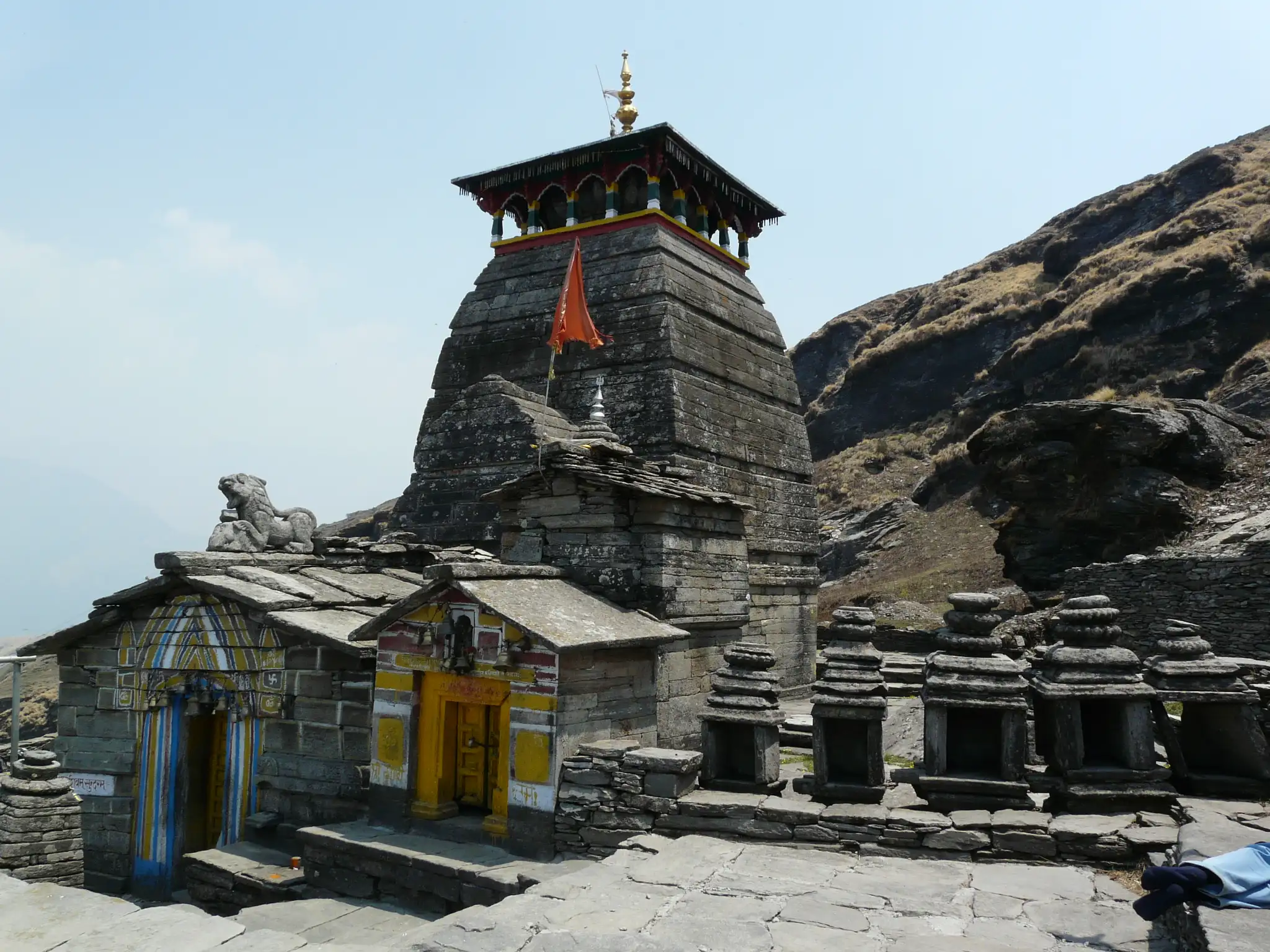 Stone shikhara of Tungnath Temple with a saffron flag, a stone lion on the roof and a row of small shrines on the paved courtyard