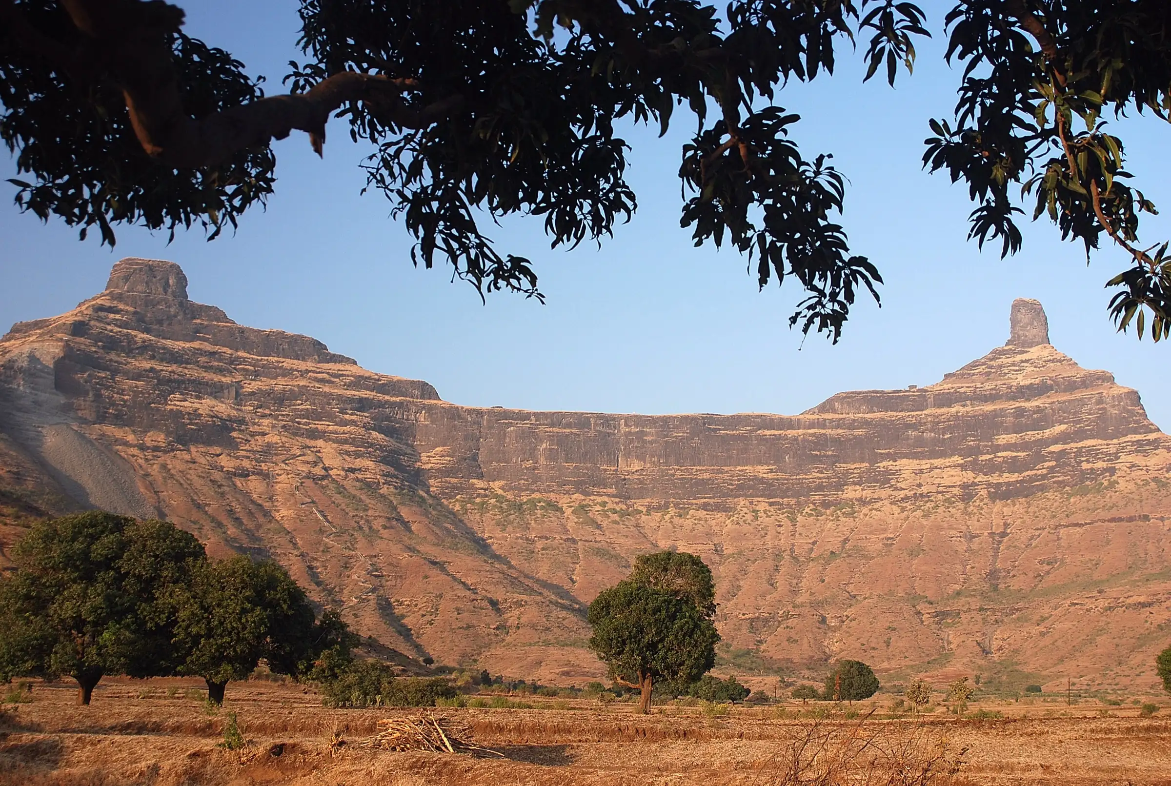 The twin rock pinnacles of Mangi and Tungi at either end of a long reddish basalt ridge, seen from fields below with mango trees