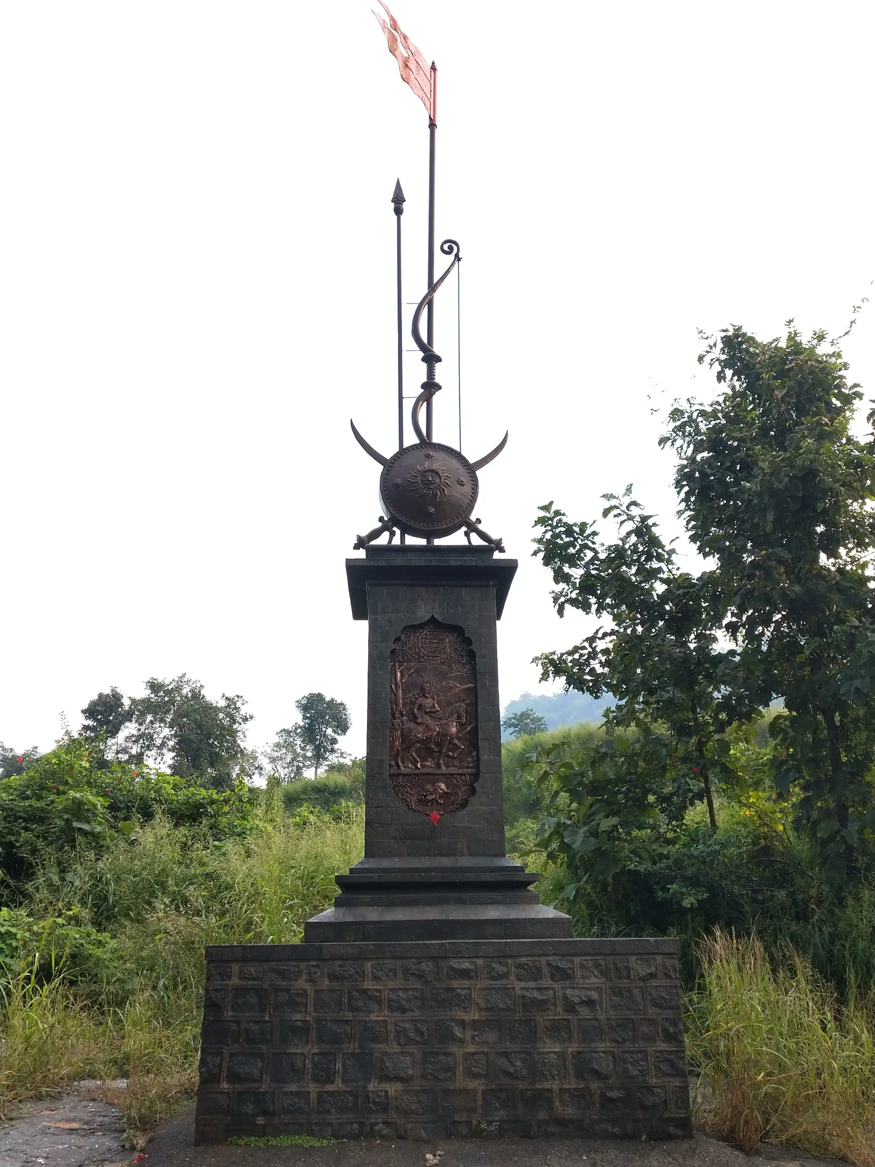 Umberkhind victory memorial: a stone pillar with a bronze relief panel, topped by a shield, spear, bow and flag, among green trees