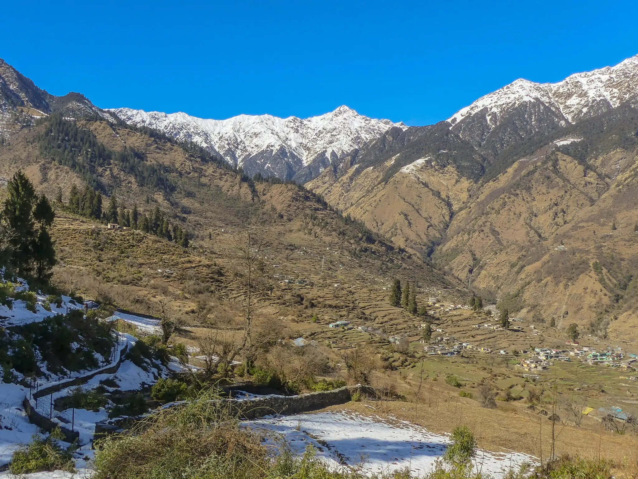 Terraced fields and scattered snow in the Urgam valley below a ridge of snow-capped peaks under a deep blue sky