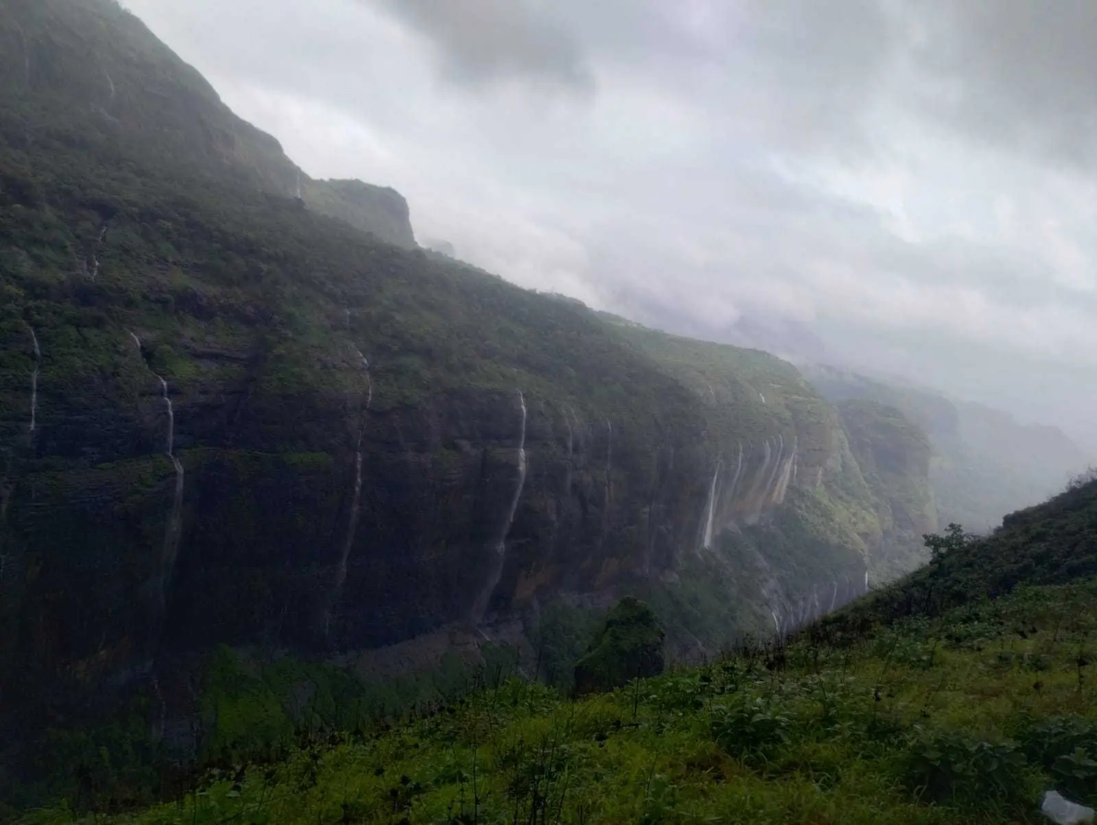 Long green cliff wall streaked with thin monsoon waterfalls above a grassy slope under low grey cloud