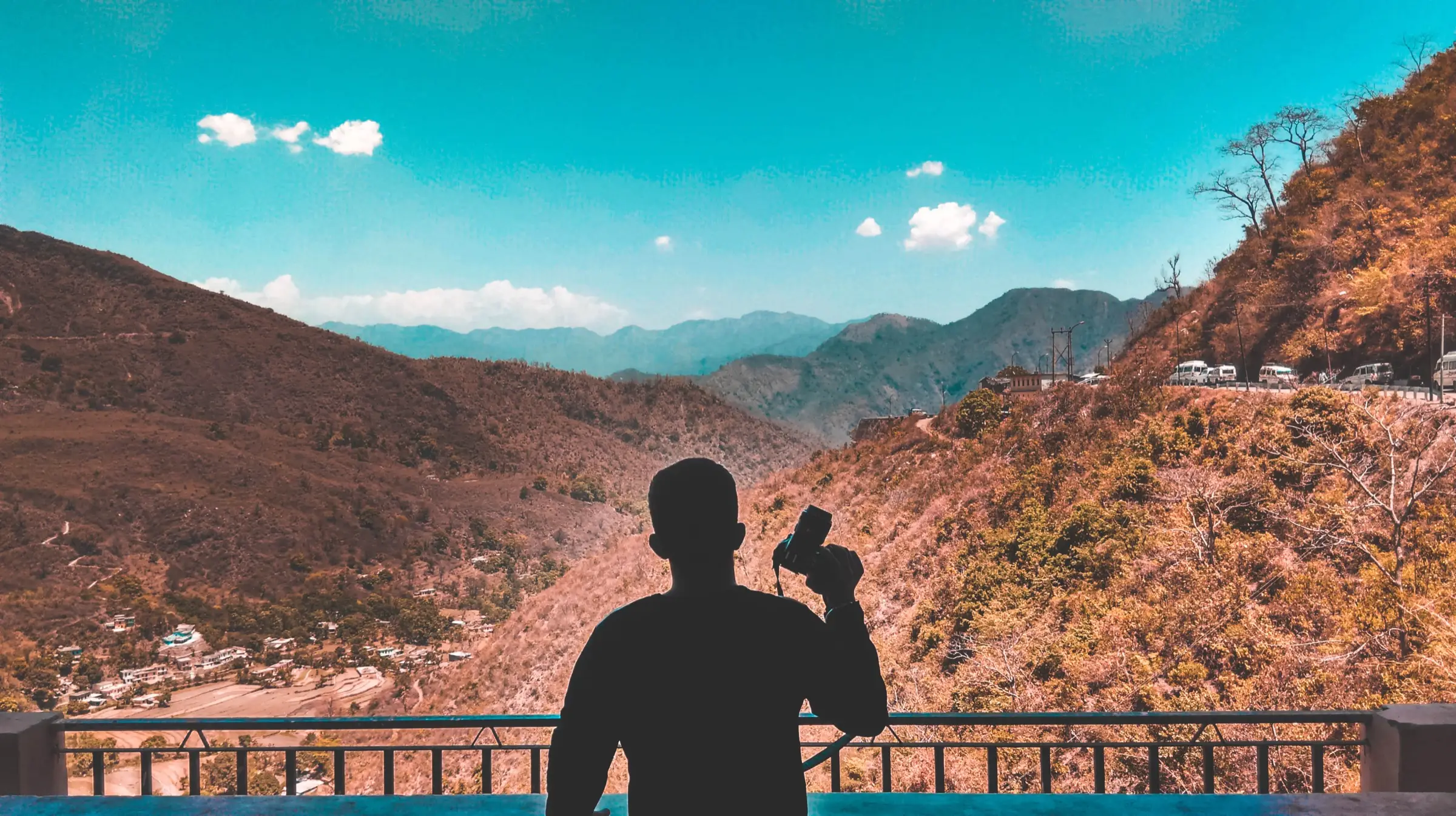 A visitor in silhouette at a railing looking over dry hills and a village in the valley on the way to Neelkanth
