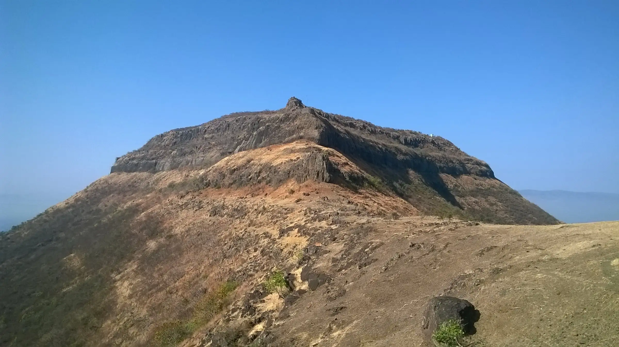 Vandan Fort's rocky scarp rising above a dry grassy ridge under a clear blue sky