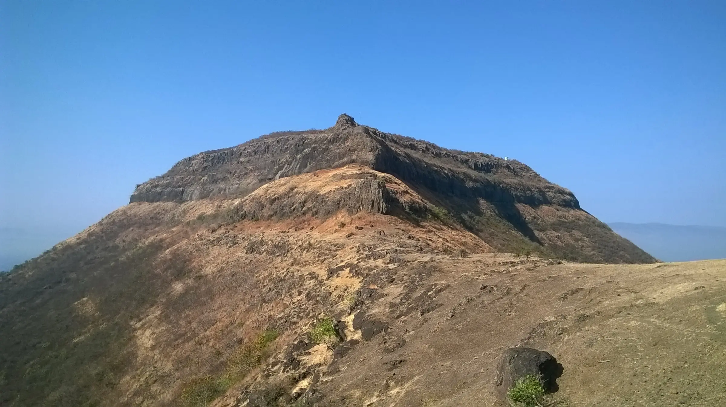 Vandan Fort's rocky scarp rising above a dry grassy ridge under a clear blue sky
