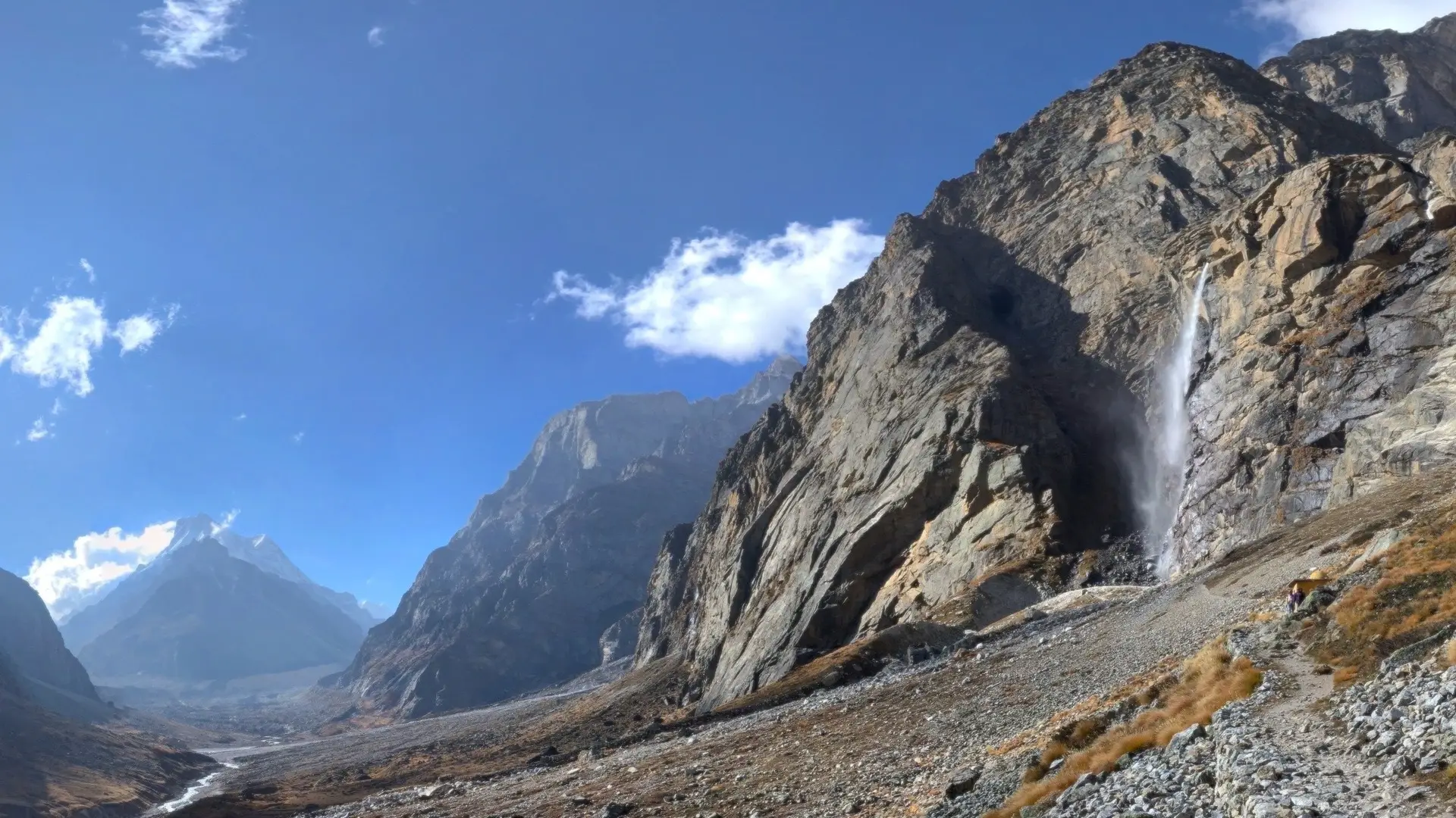 Vasudhara Falls pouring down a tall grey rock face beside the stony trail above the Alaknanda valley