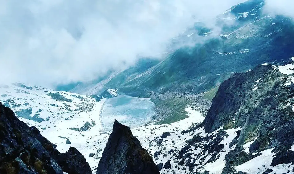 Vasuki Tal, a frozen lake in a snowy bowl below dark rock ridges, seen from above with cloud over the valley