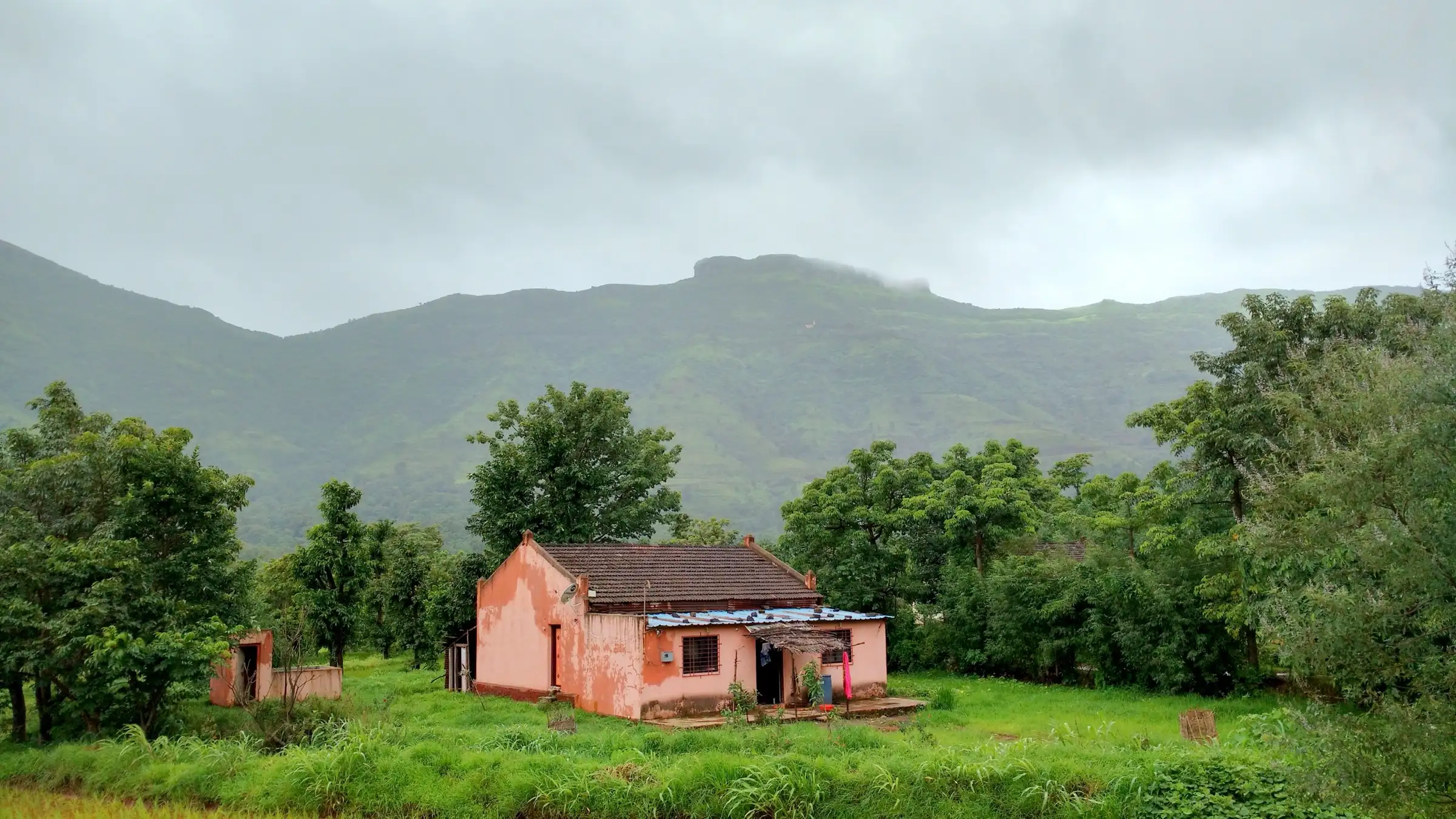 Flat-topped Kenjalgad fort under monsoon cloud above green hills, with a pink village house among trees in the foreground