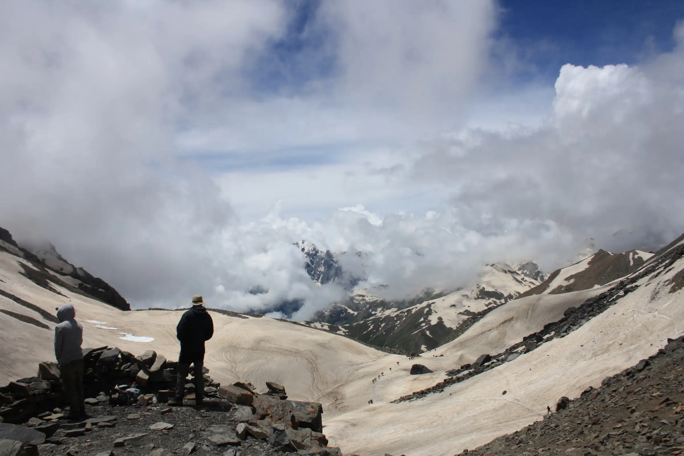 Two trekkers standing on rocks at Pin Bhaba Pass looking south over snowfields and clouded peaks