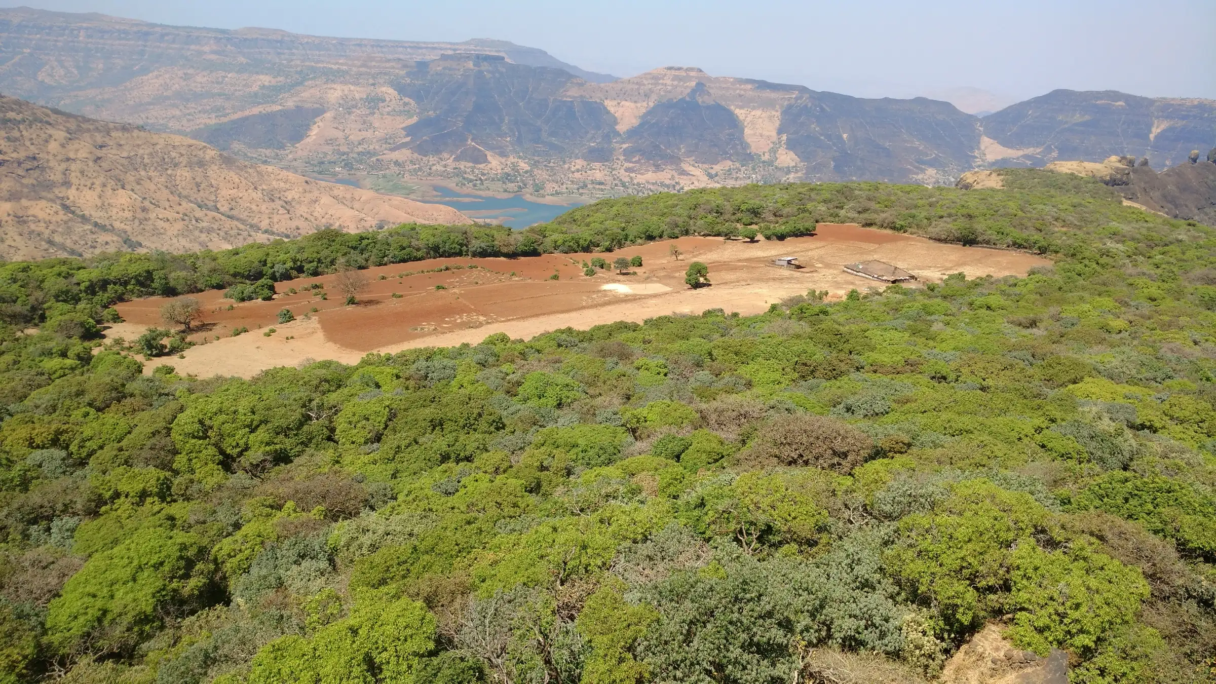 View from Kamalgad over forest and a red-soil clearing to the Dhom reservoir and the flat-topped Mahabaleshwar hills