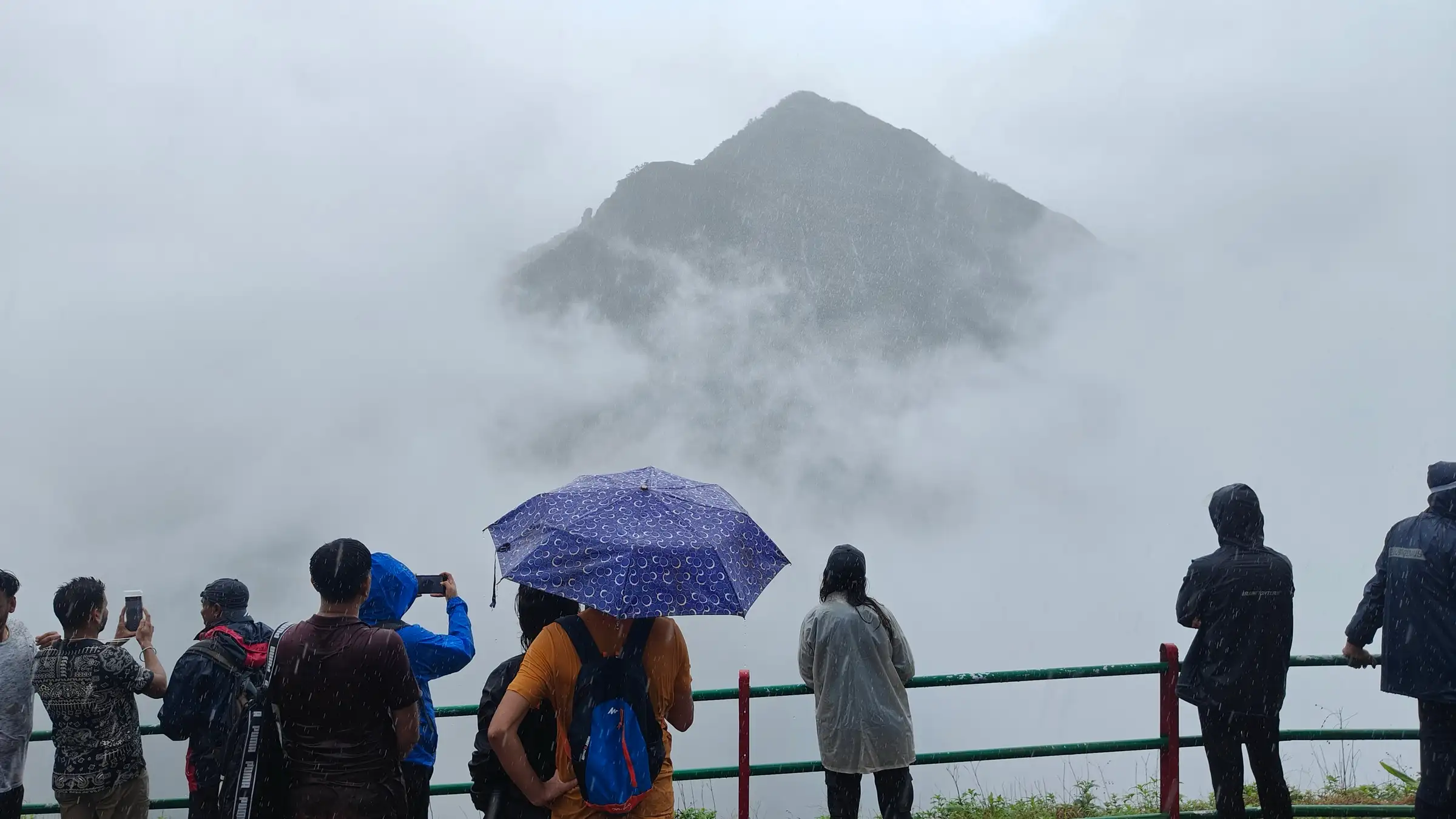 Trekkers in rain jackets and under an umbrella at a railed viewpoint looking at a pointed peak half hidden in fog
