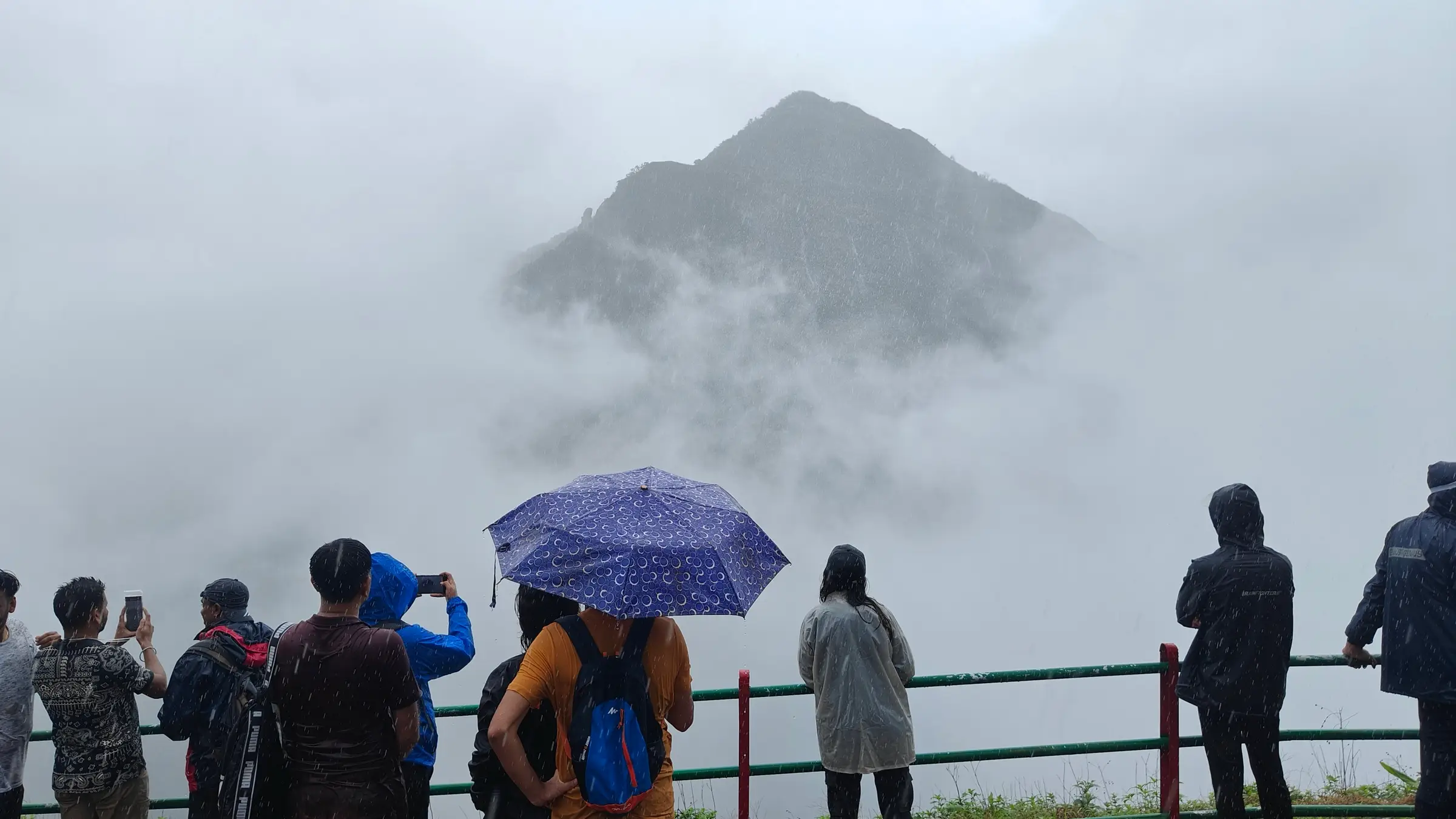 Trekkers in rain jackets and under an umbrella at a railed viewpoint looking at a pointed peak half hidden in fog