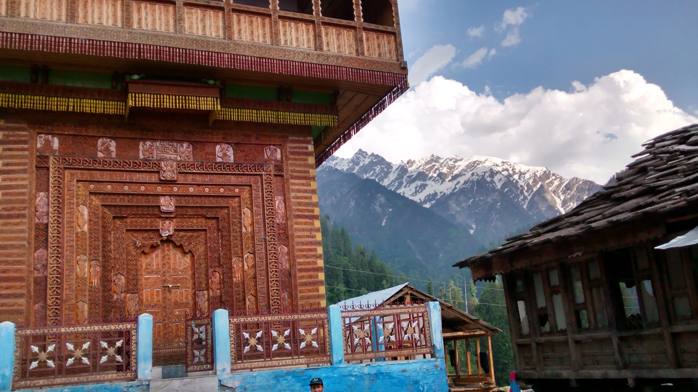 Carved wooden temple and old wooden houses in Grahan village, with snow peaks of the Parvati valley behind