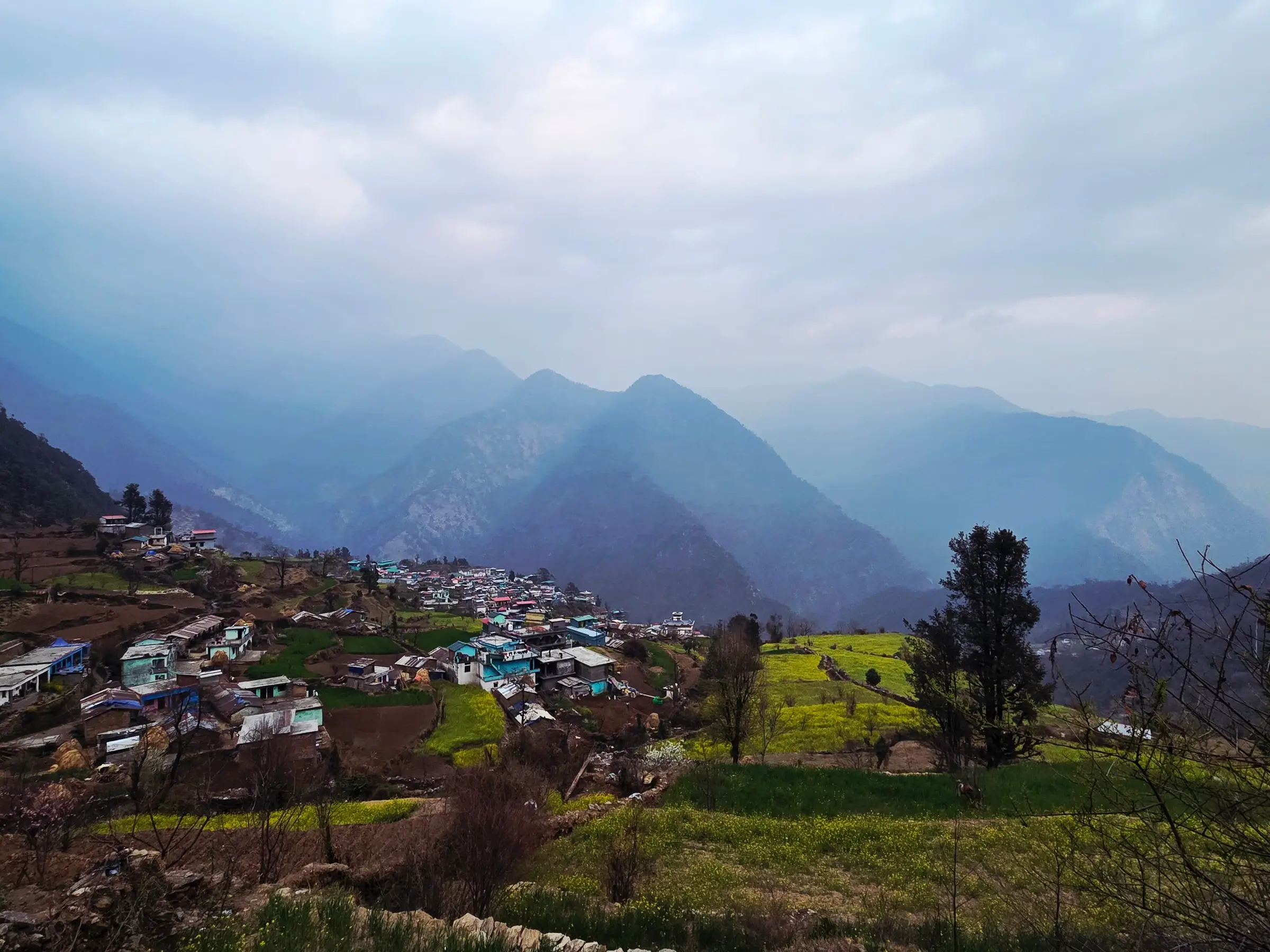 Triyuginarayan village spread over green terraced fields, with layered blue mountain ridges of the Kedar valley in the haze beyond