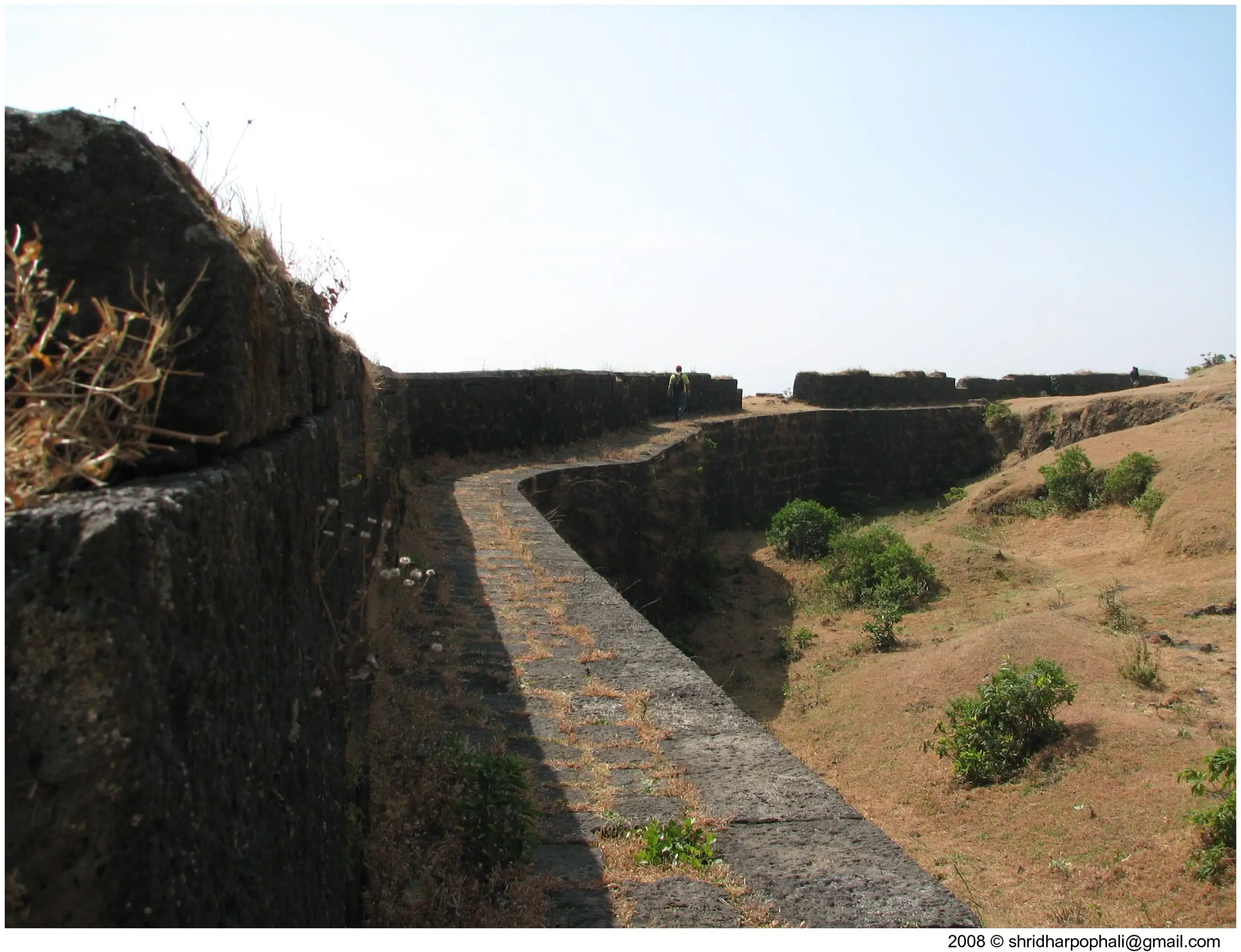 A trekker walking along the winding stone fortification wall of Visapur Fort across dry grass on the plateau