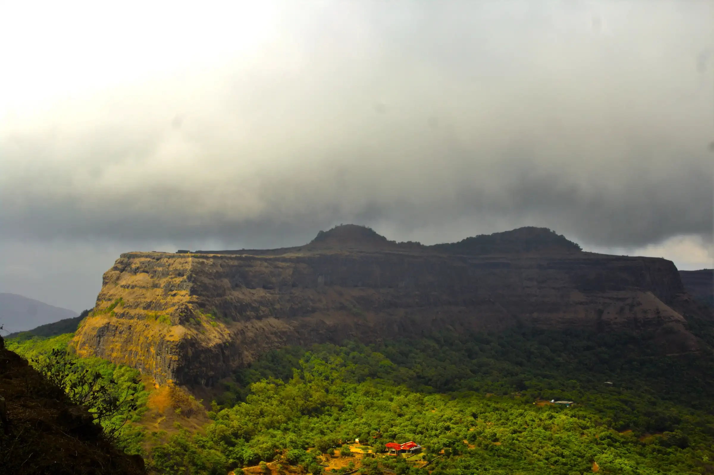 Visapur Fort's long, sheer-sided basalt plateau under monsoon clouds, seen from Lohagad above a green forested valley