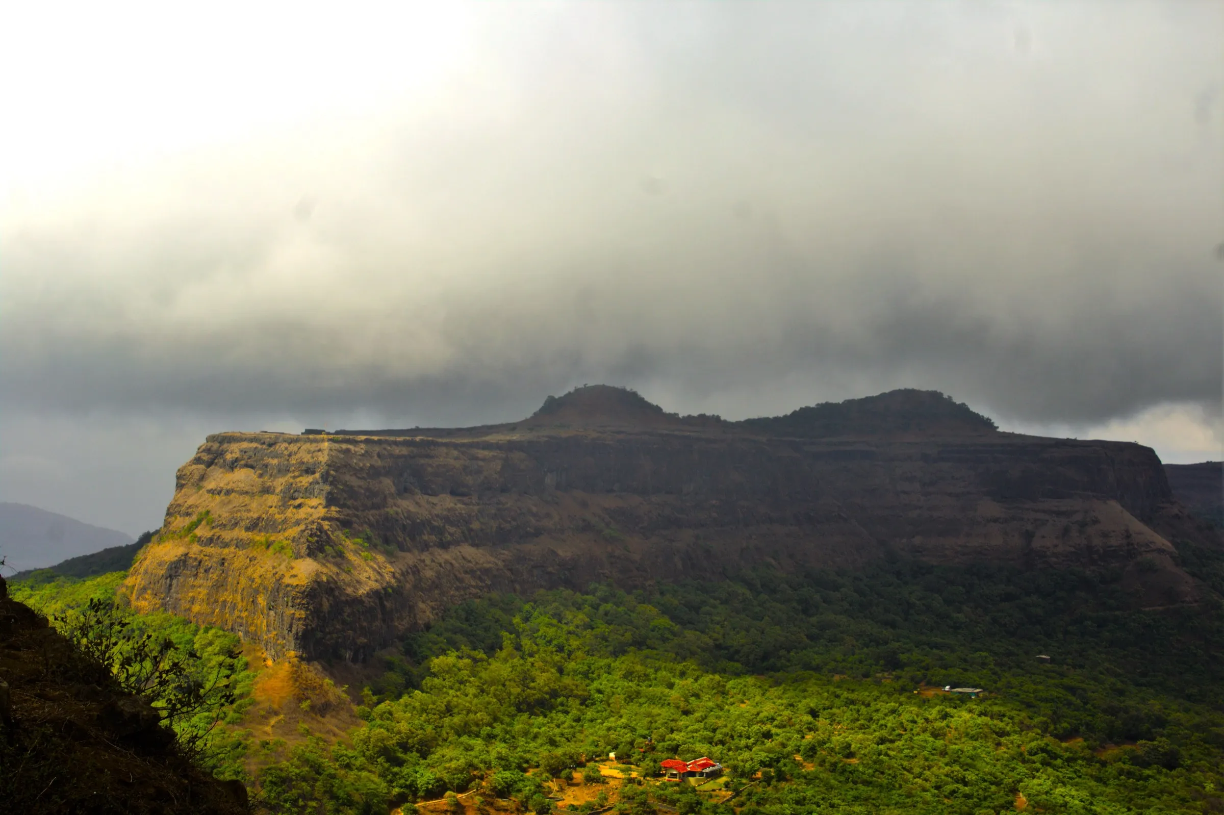 Visapur Fort's long, sheer-sided basalt plateau under monsoon clouds, seen from Lohagad above a green forested valley