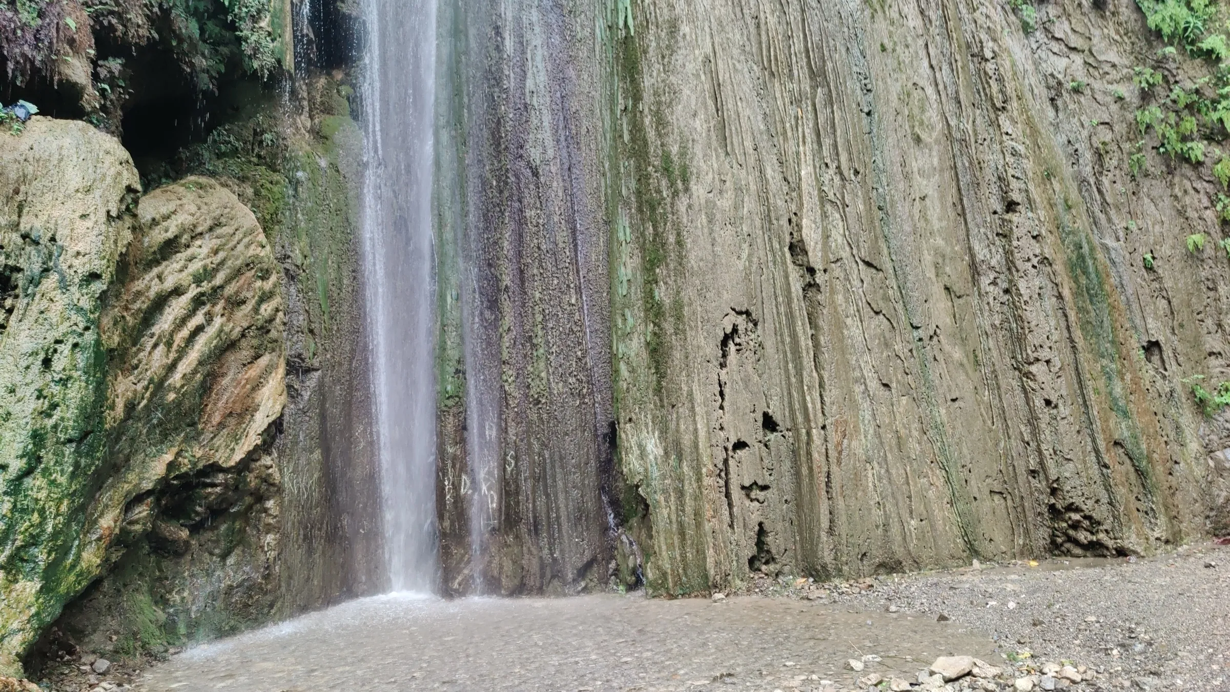 Patna Waterfall pouring as a thin white column down a tall, grooved limestone cliff onto a gravel pool floor