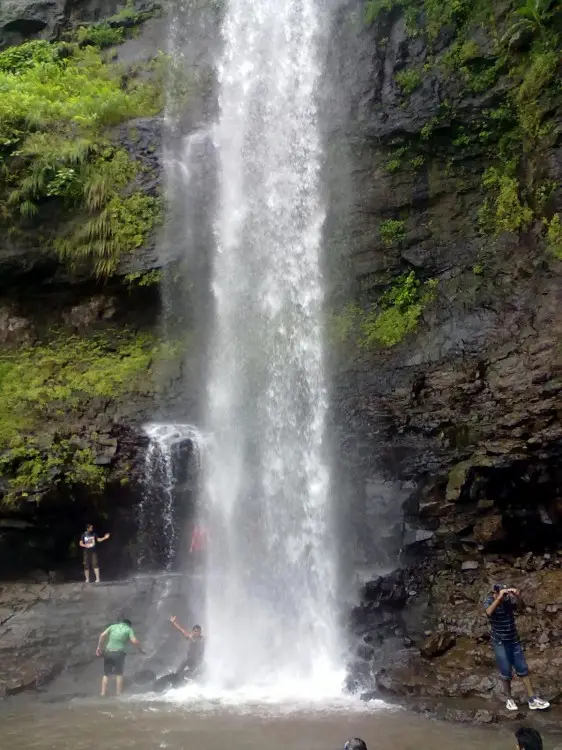 Tall monsoon waterfall at Siddhagad pouring down a dark basalt cliff with visitors at its base