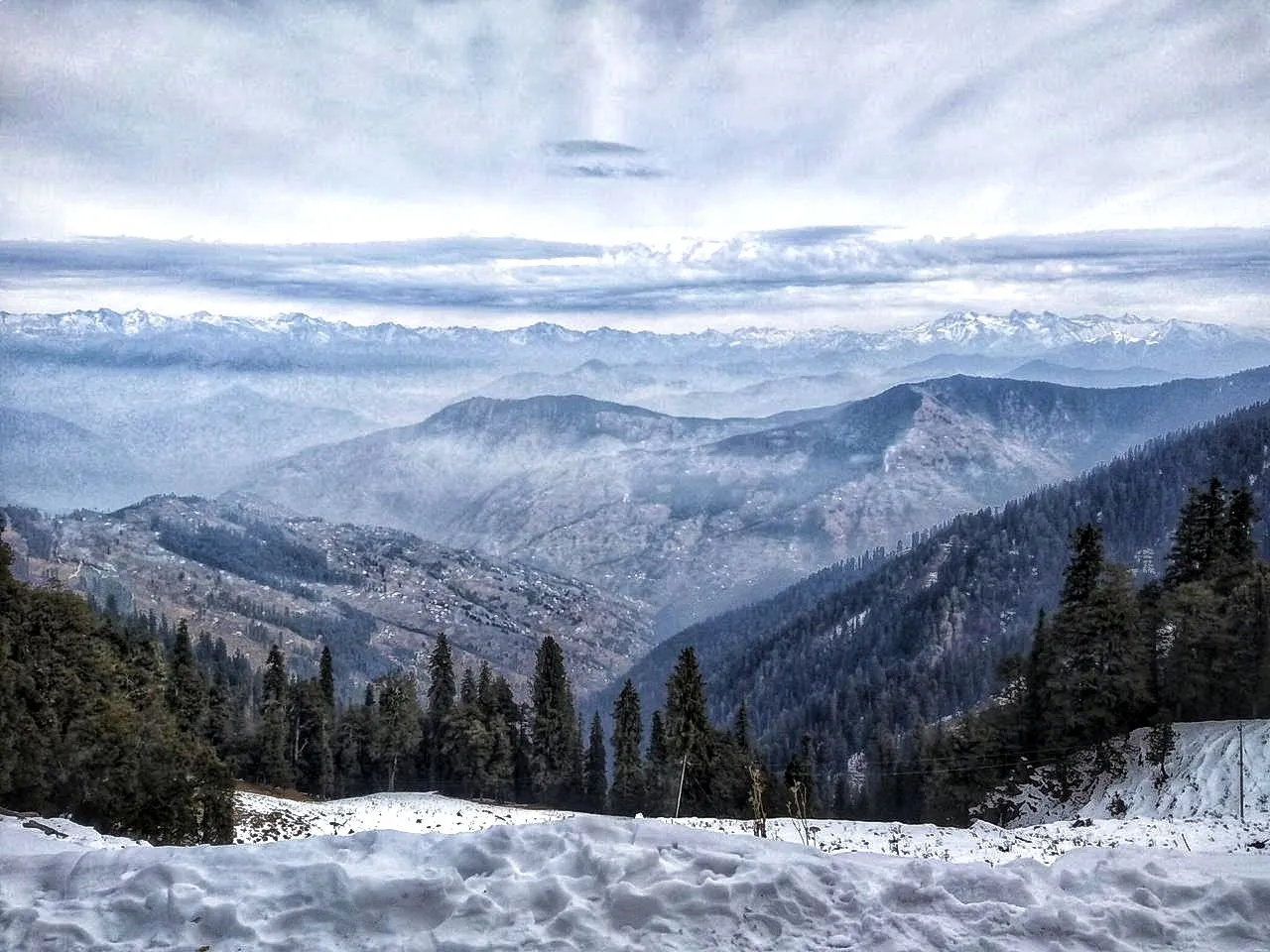 Snow on the road edge and dark pine forest above folded valleys, with the snow ranges on the horizon, seen on the way to Hatu Peak