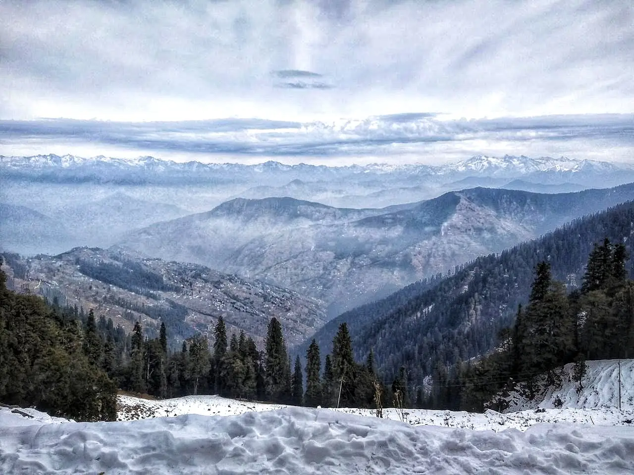 Snow on the road edge and dark pine forest above folded valleys, with the snow ranges on the horizon, seen on the way to Hatu Peak
