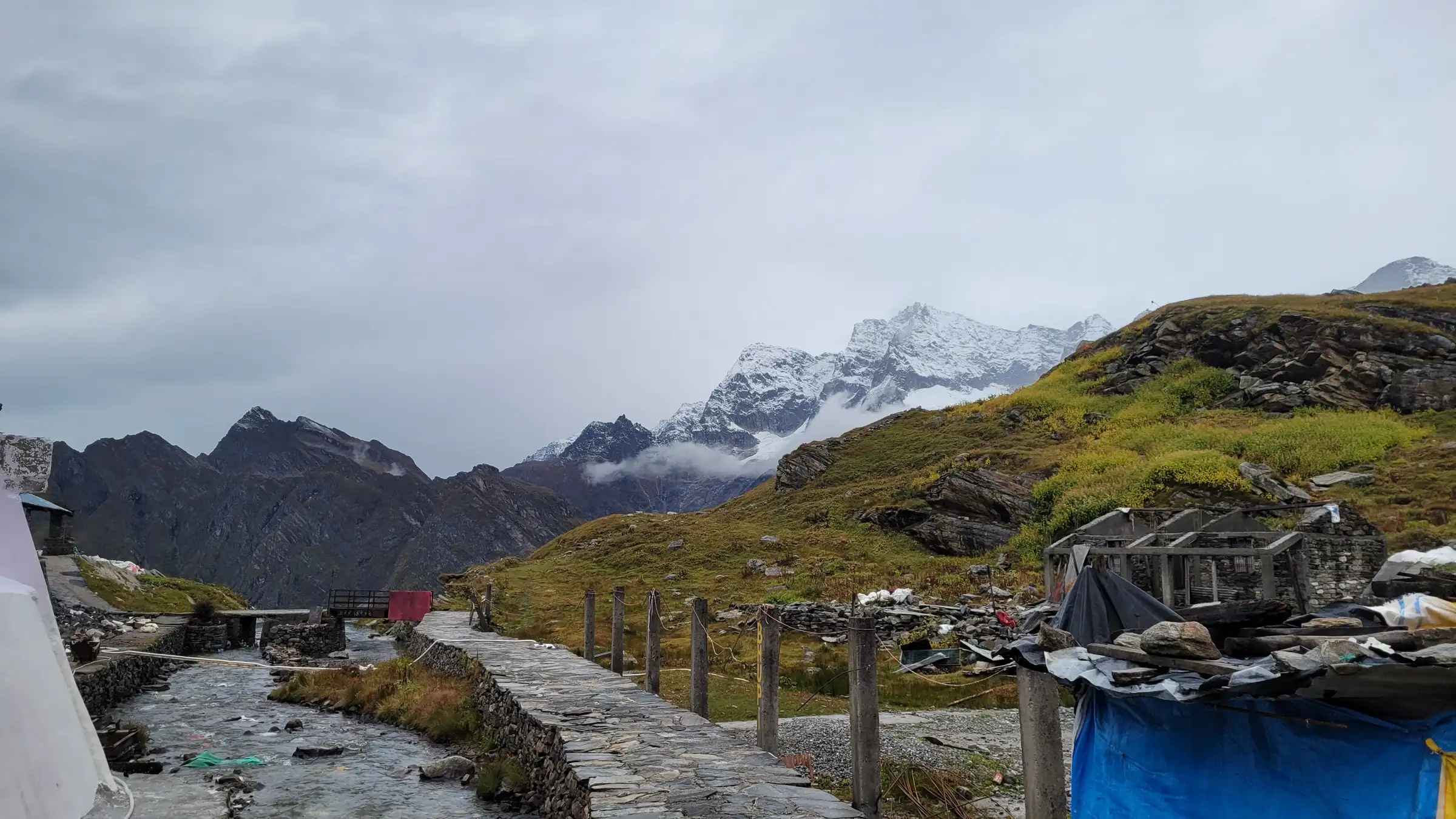 Stone-paved pilgrim path beside a stream and wooden fence on the way to Hemkund Sahib, with snow peaks behind