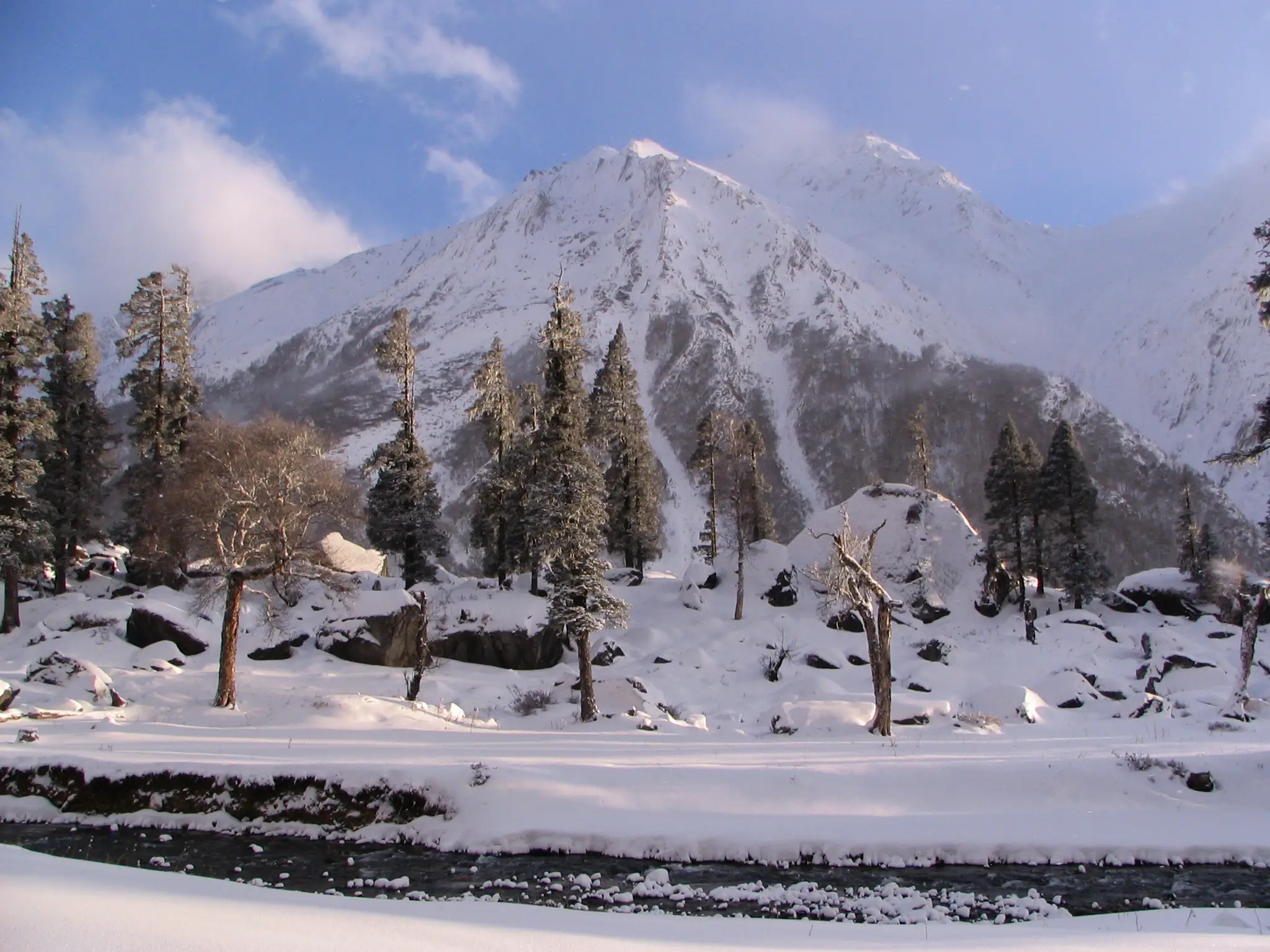 Har Ki Dun under deep winter snow with conifers and a dark stream in front of a white mountain