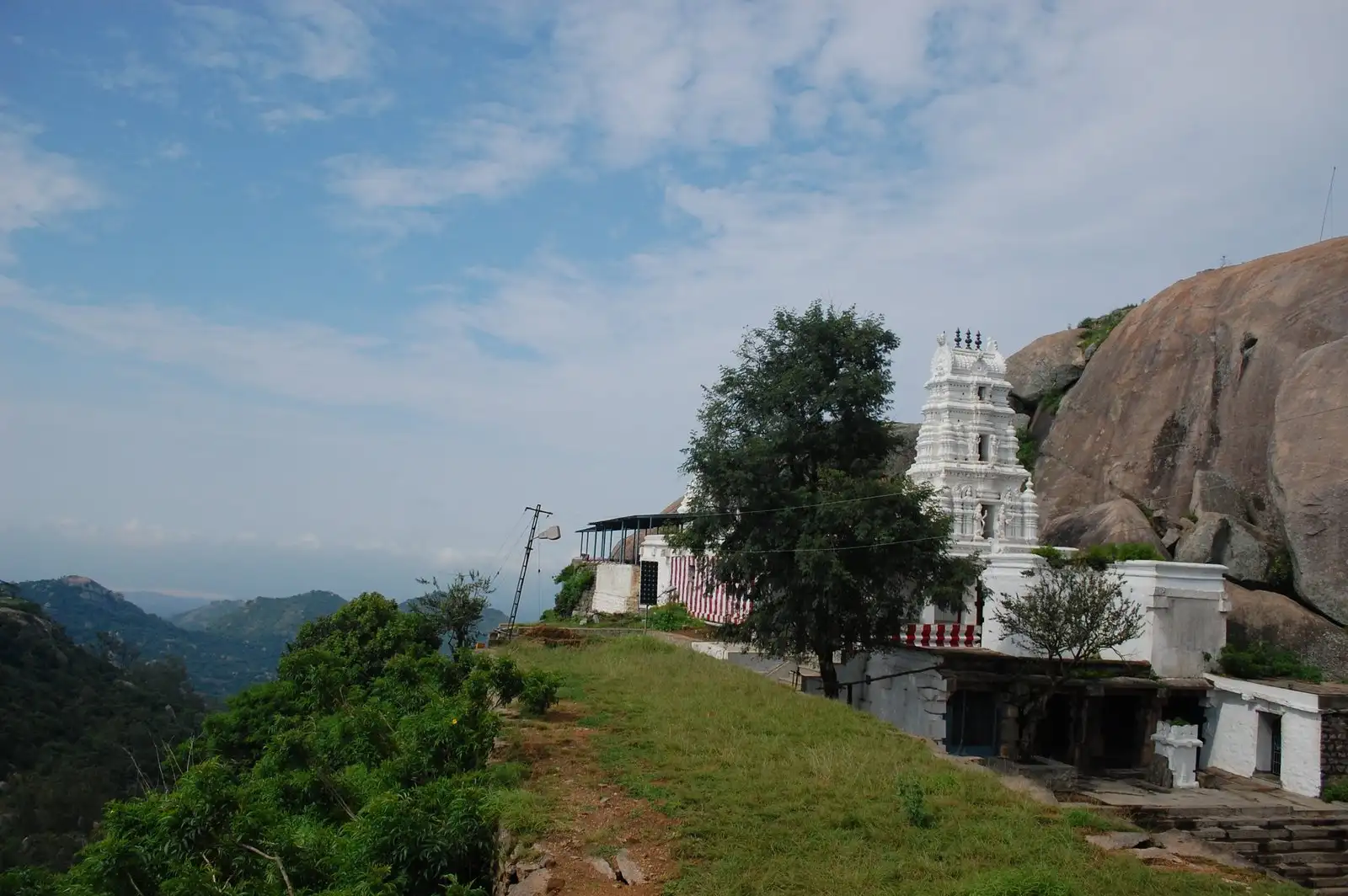 White gopuram of the Yoga Narasimha Swamy temple atop Devarayanadurga hill, set against a huge granite rock with forested hills behind