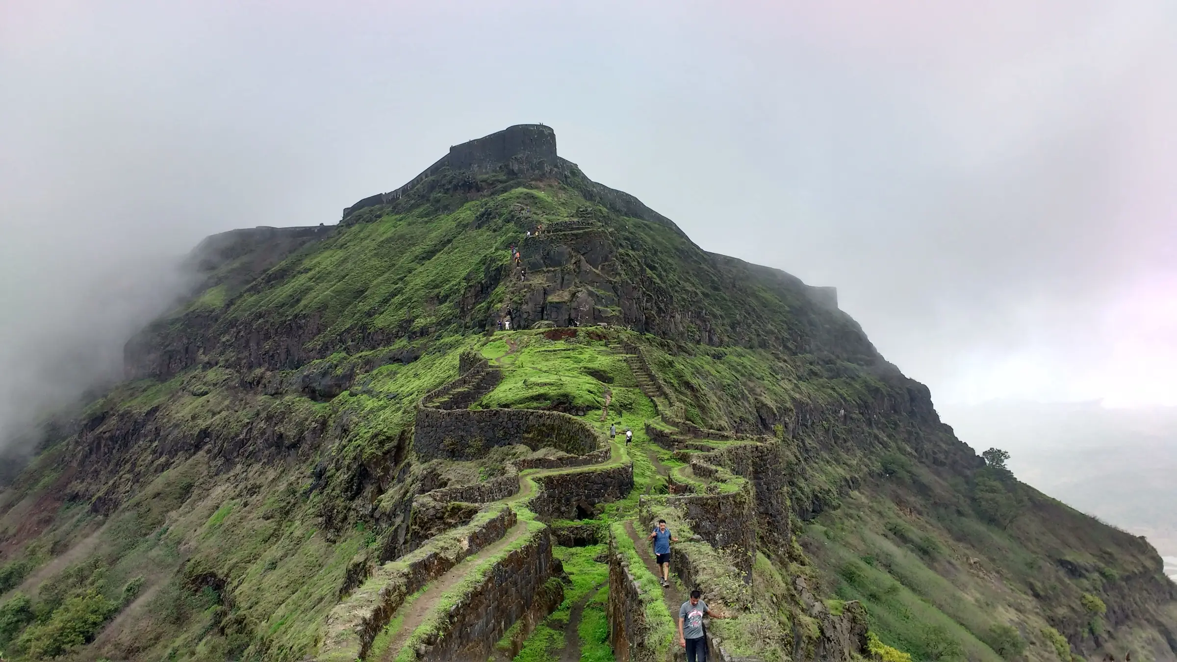 Trekkers walking along the stone fortification walls of Zunjar Machi towards the misty top of Torna Fort